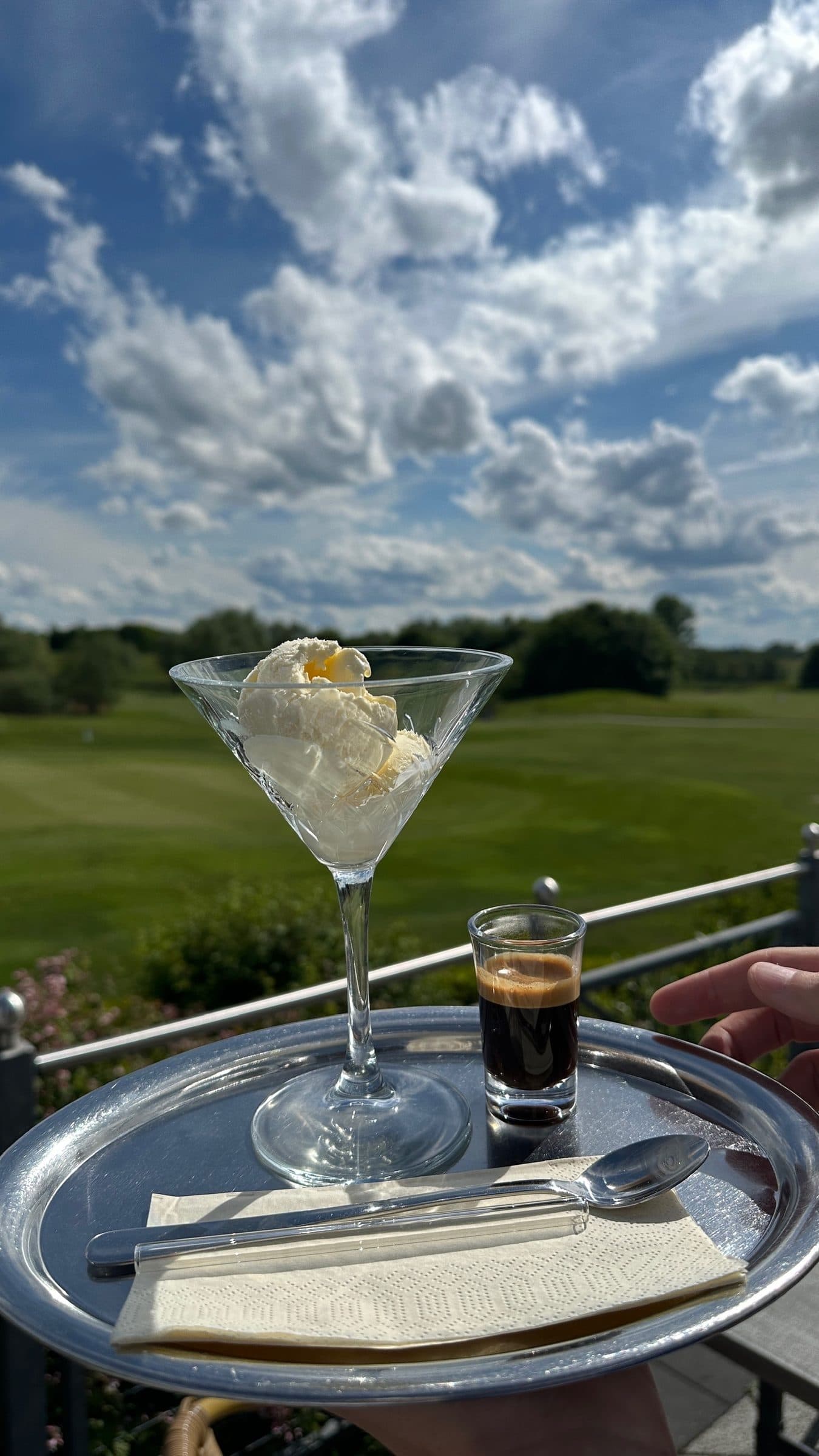 Vanilla ice cream in a glass and an espresso on a tray, the fairway and summer clouds behind