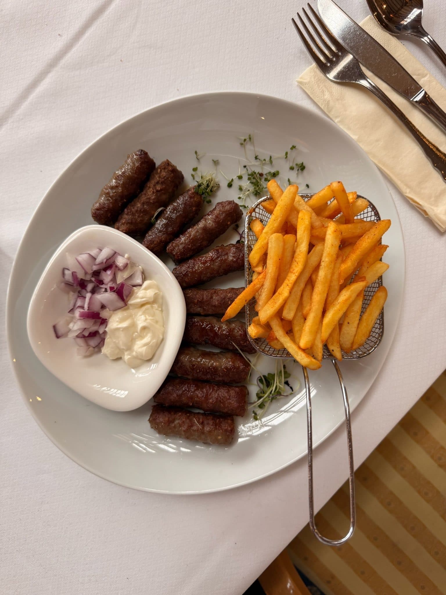 Ćevapi mit Pommes, roten Zwiebeln und Kajmak auf einem weißen Teller