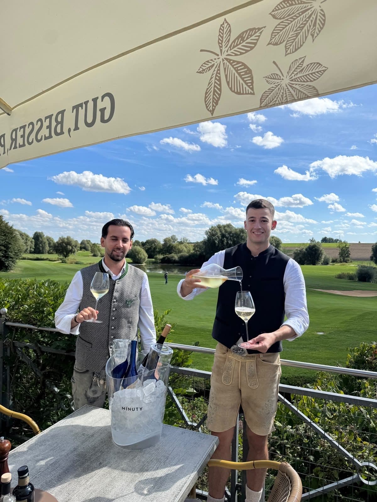Zwei Männer in Trachtenweste schenken auf der Terrasse Wein aus, dahinter der Golfplatz