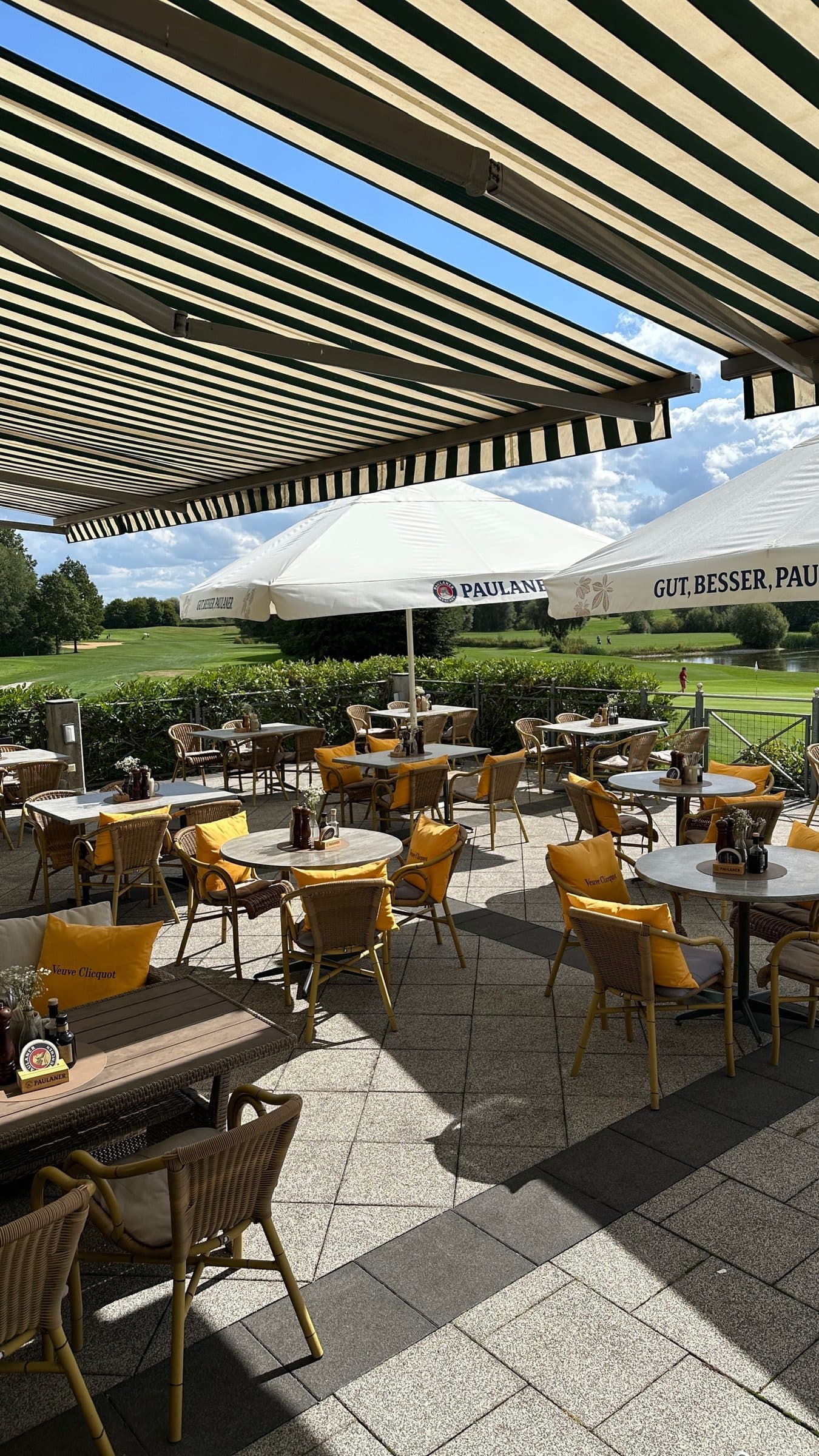 View from beneath the awning across the terrace with wicker chairs and Paulaner parasols, the fairways beyond