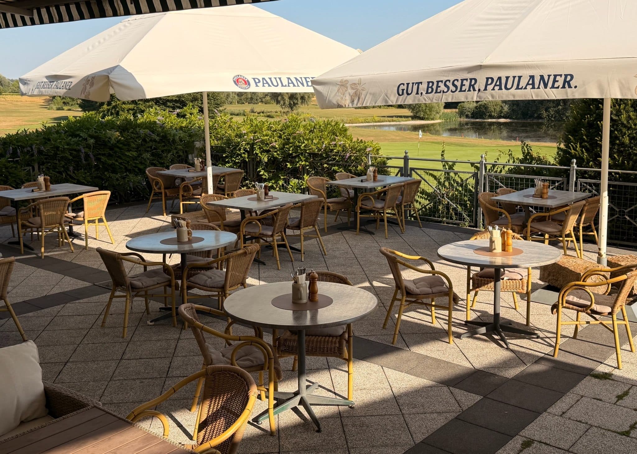 Die Terrasse mit Paulaner-Sonnenschirmen und Korbstühlen, dahinter der Golfplatz